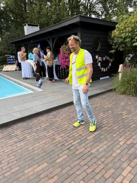 A lively outdoor gathering is underway at 't Jannendorp, Leusden, featuring a man in a bright yellow vest smiling while playing a game near a swimming pool